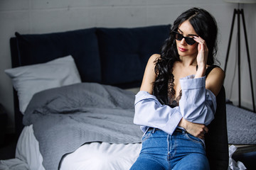 portrait of a beautiful brunette in sunglasses, blue shirt, blue jeans and interior in room