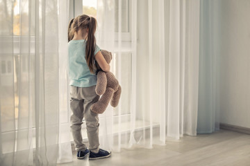 Lonely little girl with teddy bear near window. Autism concept