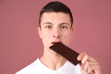 Young man with acne problem eating chocolate bar on color background. Skin allergy