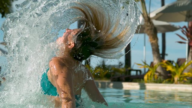 SLOW MOTION, CLOSE UP: Brunette girl in luxury pool flips her hair and sprays pool water behind her. Glistening drops of water are sent flying after cheerful woman whips her head out of awesome pool.