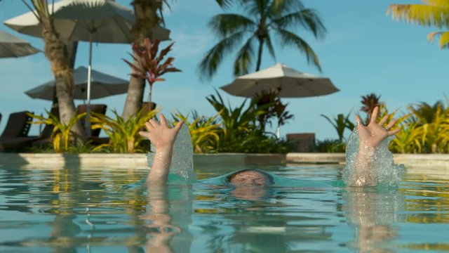 SLOW MOTION, CLOSE UP. REVERSED: Excited girl with long brown hair jumps out of stunning exotic pool. Happy girl jumping and splashing glassy water in high end oceanfront pool in breathtaking Fiji.