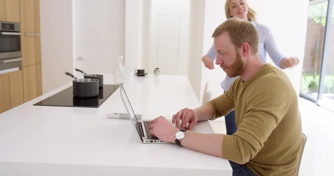 Portrait of attractive couple as man works from home as partner comes and happily chats to her about what she’s doing, in contemporary kitchen, in slow motion