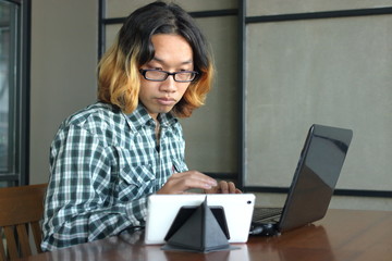 Relaxed young Asian hipster man working against laptop and tablet in his office.