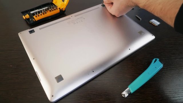 The master fastens the back cover of the laptop with screws. Close-up hands tighten the screw using a screwdriver.