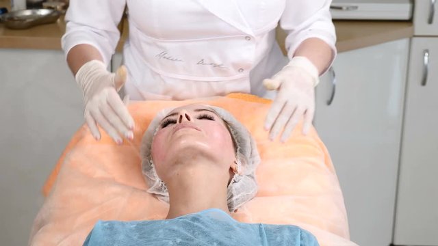 Beautician in gloves massages young female face before applying beauty cosmetic mask. Young pretty female client gets facial beauty procedure.4k