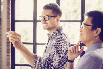 Closeup of smiling young business man writing on whiteboard and his senior Asian boss with loft...