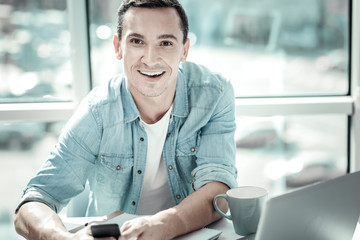 Work with pleasure. Satisfied gay pleasant man sitting by the table smiling and holding his cellphone.