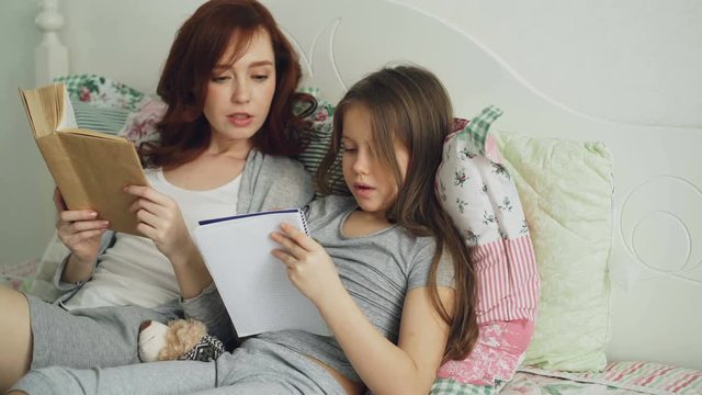 Careful mother helping her little cute daughter with homework for elementary school. Loving mom reading a book and girl writing notes in copybook while sitting together on bed at home