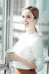 Be the best. Confident pretty stylish woman standing in the bright room near the window holding a cup and smiling.