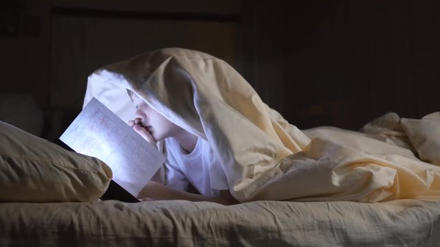 A child reads a book under blankets with a flashlight at night. Enthusiastic boy