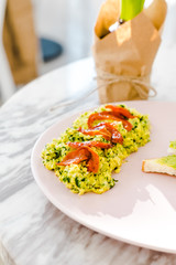 Spinach scrambled eggs with avocado toast on pastel pink plate. White marble table at the cafe. Sunny morning breakfast. Food photography concept