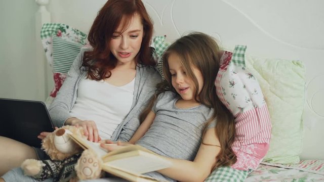 Little cute daughter with young loving mother watching funny pictures and laughing while reading fairy tale book sitting together on cozy bed at home