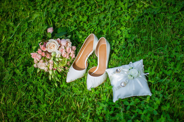 Wedding accessories on green grass in summer. Two gold wedding rings on a white pillow. Wedding shoes for women. Bouquet of flowers. 