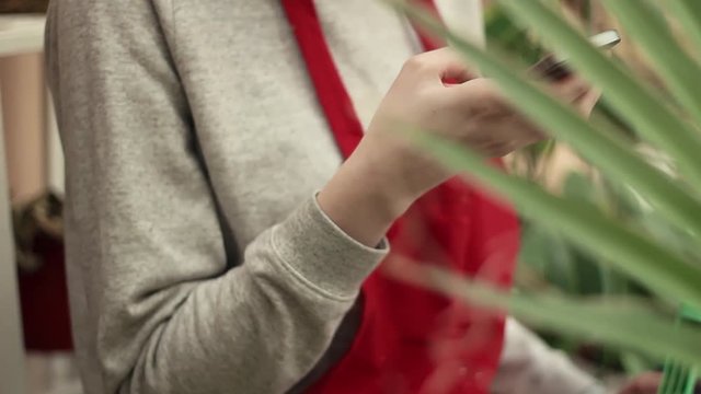 Hands of young woman gardener in red apron using smartphone in greenhouse.