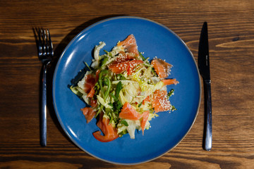 A horizontal image of salad with salmon, lettuce, arugula, tomatoes, sesame seeds. Served on a bright blue plate.