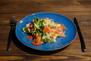 A horizontal image of salad with salmon, lettuce, arugula, tomatoes, sesame seeds. Served on a bright blue plate.