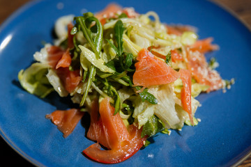 A horizontal image of salad with salmon, lettuce, arugula, tomatoes, sesame seeds. Served on a bright blue plate.