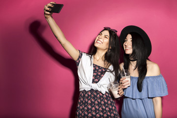 Two caucasian brunette hipster woman in casual stylish outfit having fun making selfie on their phone. They standing on a bright pink background. Cheerful, happy emotions