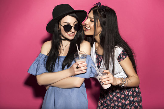 Two caucasian brunette hipster woman in casual stylish outfit having fun drinking milkshakes with straws. They standing on a bright pink background. They gossip with a cheerful, happy emotions