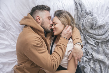 Stylish hipster couple of man and woman sitting on bed in their home in cozy casual clothes. They hug and love each other and smile happily, resting together. Space for text