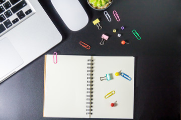 Obraz premium Top view of office table or desk with blank notebook, laptop, colorful equipment and smart phone on black background with copy space for text.