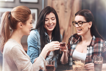 To our friendship. Joyful cheerful female friends sitting together and cheering with shots while drinking to their friendship