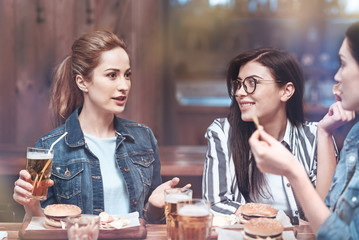 Interesting story. Beautiful positive good looking woman sitting at the table and drinking beer while telling a story to her friends