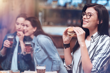 Favourite food. Happy delighted positive woman smiling and holding a burger while eating in the pub with her friends
