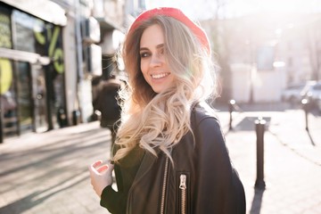 Lovely friendly woman with long blond hair wears red hat and leather jacket enjoys the sun and walks on street in sunlight with beautiful smile