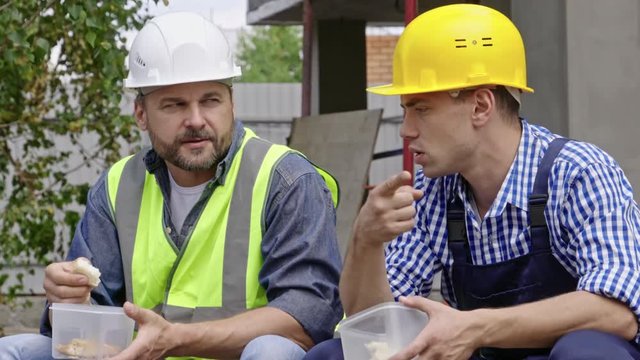 Medium shot of two male engineers sitting at construction site, having friendly conversation and eating lunch during break from work