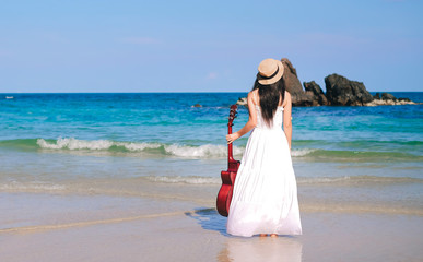 Fototapeta na wymiar woman traveler enjoying for view of the beautiful sea on her holiday. Lady tourist standing and holding a guitar on the beach.