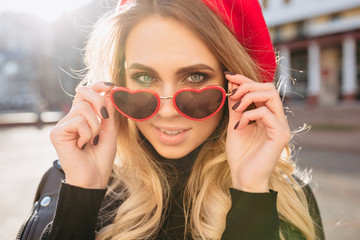 Naklejka premium Close-up portrait of attractive blond female model dressed red hat and stylish glasses in sunlight. Photo of happy caucasian girl in sunglasses posing outside.