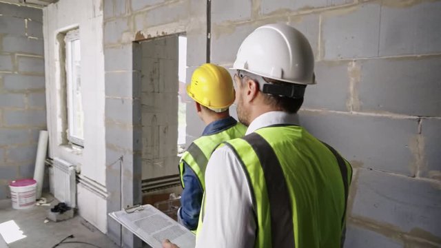 Two men wearing hard hats and safety vests walking inside building under construction, one showing work progress to another one, follow shot