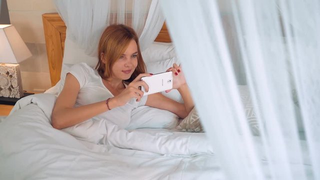 Young caucasian woman using smartphone while lying in white canopy bed