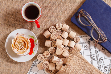 Board game lotto on sackcloth. Wooden lotto barrels in bag and game cards with notebook, glasses, cup of coffee and homemade cookie on plate