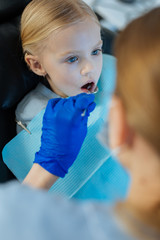 Skilled checkup. Pleasant petite girl sitting in a dentist chair and undergoing an oral cavity checkup while a female dentist using a mouth mirror