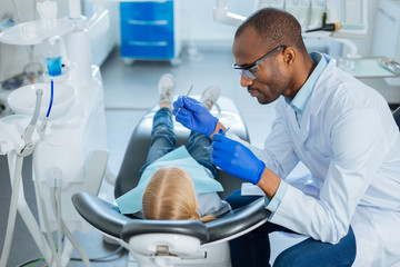 Fototapeta premium Best teeth care. Pleasant caring male dentist using a dental probe and a mouth mirror and checking the teeth of a little girl lying in the chair