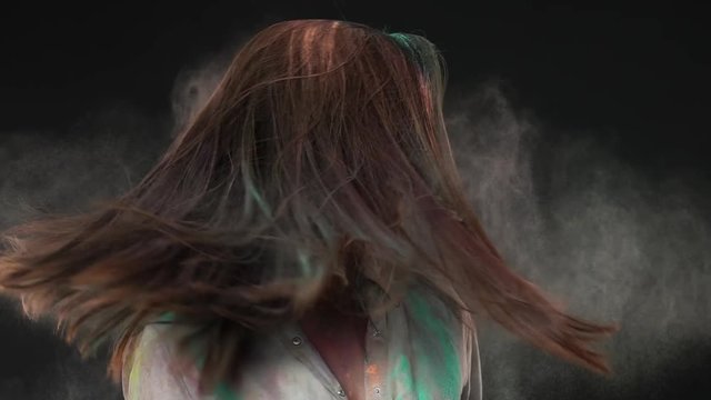 Portrait of caucasian woman smiling and shaking head with multicolor dust in her brown hair on holi carnival of colors, isolated over dark gray background. Indian culture and tradition