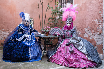 Fototapeta na wymiar Two masked ladies in blue and pink costumes sitting at a table in a courtyard with an old terracotta wall behind them at Venice Carnival (Carnivale di Venezia)