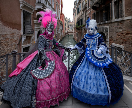 Two women in pink and blue costumes with fans and ornate painted feathered masks at Venice Carnival. Women are standing on a bridge with canal in background.
