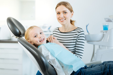 Precious support. Charming little girl and her mother posing for the camera while the girl sitting in the dentist chair and her mother supporting her before the treatment
