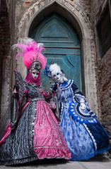 Fototapeta na wymiar Two women in masks and ornate blue and pink costumes standing in front of an old blue door at the end of an alley in Venice during the carnival (Carnivale di Venezia).
