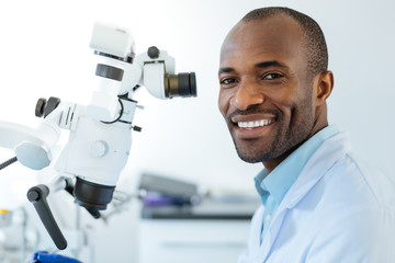 Happy user. Charming upbeat dentist posing for the camera and smiling brightly while using a microscope at work and conducting checkup