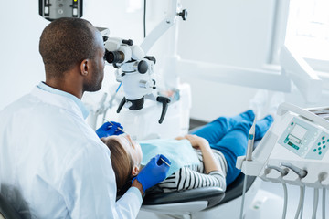Naklejka na ściany i meble Attentive to details. Professional young male dentist using a microscope and looking for cracks in his patients teeth while she lying in the chair peacefully