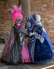 Fototapeta na wymiar Two women in masks and ornate blue and pink costumes and decorative fans, standing against an old brick wall. 