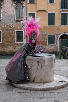 Woman in pink and black hand made costume with fan and ornate painted feathered mask at Venice Carnival. Woman looks straight to camera with canal in background.
