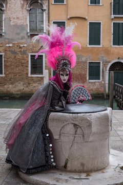 Woman in pink and black hand made costume with fan and ornate painted feathered mask at Venice Carnival. Woman looks straight to camera with canal in background.