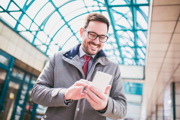 Fototapeta premium Businessman using mobile phone outside of office buildings in the background. Young caucasian man holding smartphone for business work.