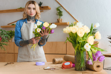 young florist in apron holding beautiful tulips and looking at camera in flower shop