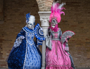 Naklejka premium Masked women in ornate blue and pink costumes standing next to stone column in a courtyard during the carnival (Carnival di Venezia)